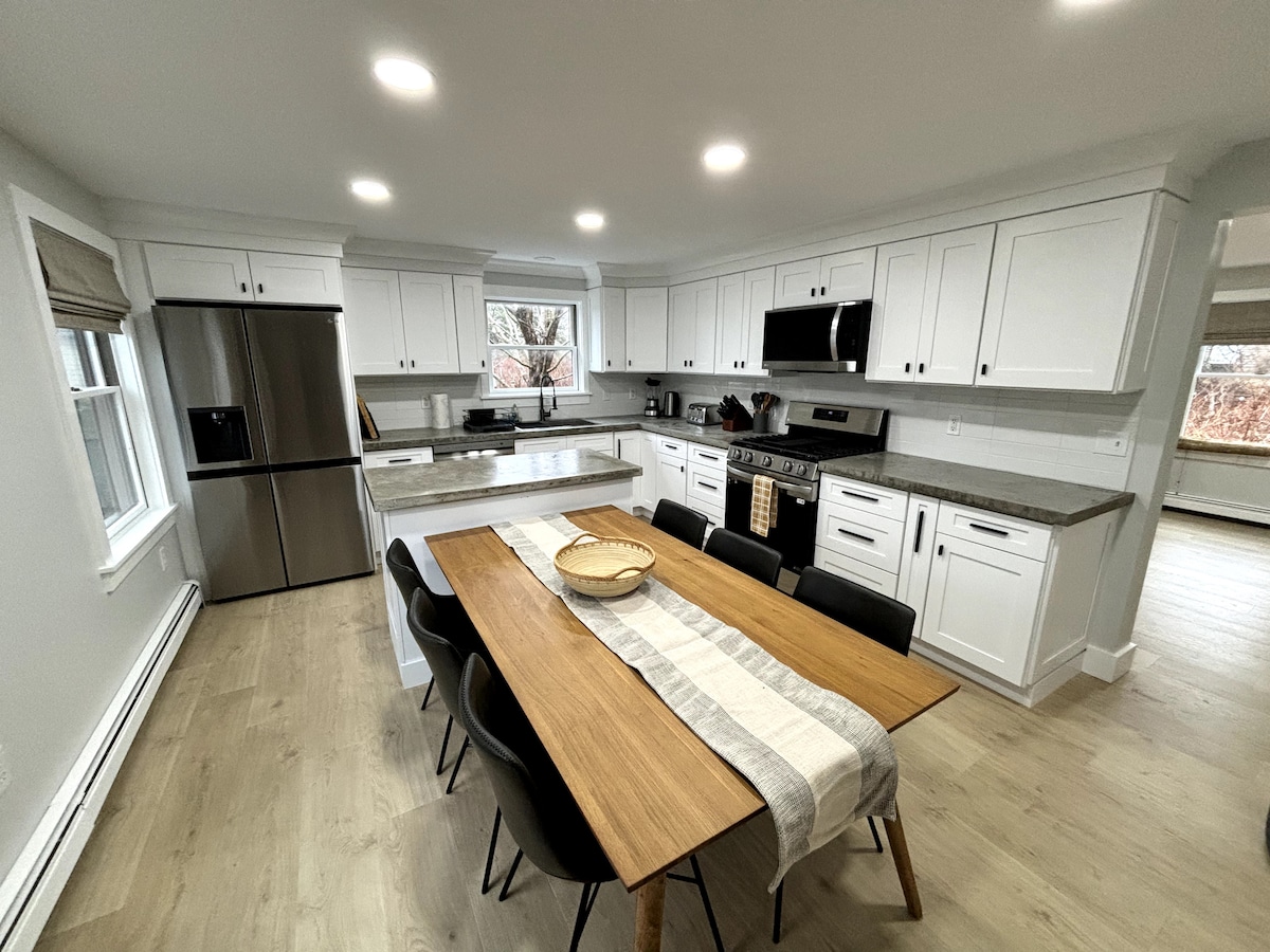 A modern kitchen is presented with white cabinetry and stainless steel appliances. A central island is paired with a wooden dining table, surrounded by six black chairs. Ample natural light enters through the windows, highlighting the sleek countertops and contemporary fixtures.