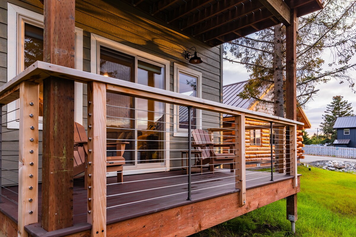 A wooden balcony features two sturdy Adirondack chairs positioned for relaxation. Natural light filters across the deck, highlighting the clean lines of the railing and the texture of the wood. In the background, a rustic log cabin complements the surroundings.