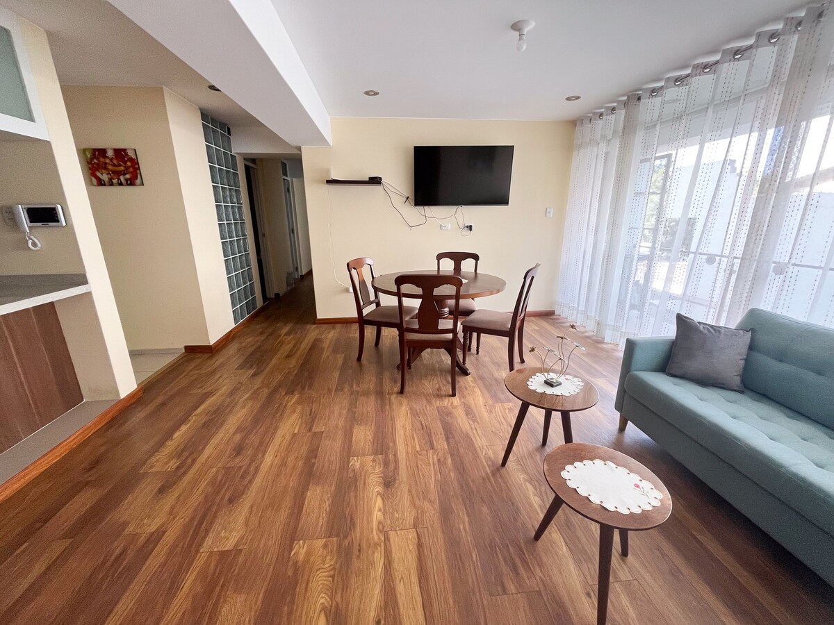 A spacious living area is presented with wooden flooring. A round dining table with four chairs is positioned near a large window, allowing natural light to brighten the space. A cozy seating arrangement, including a green couch and two small tables, enhances the inviting ambiance.