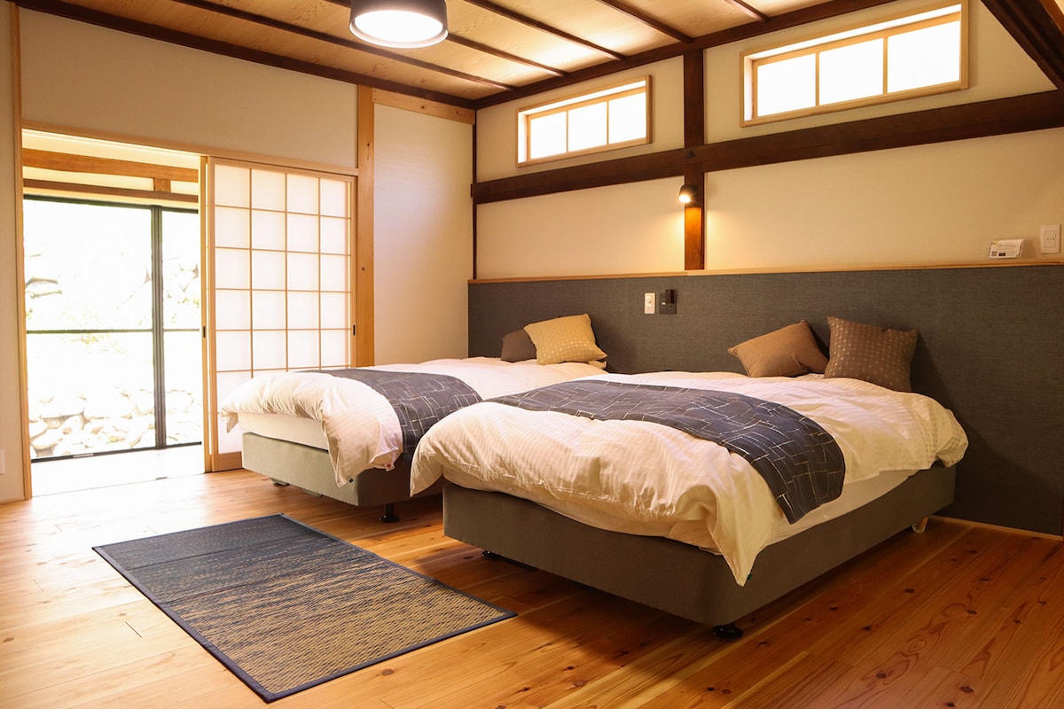The sleeping area contains two spacious beds with light bedding and decorative pillows, set against a backdrop of natural wooden accents. Soft light filters in through the windows and shoji screens, creating a serene atmosphere in the room.
