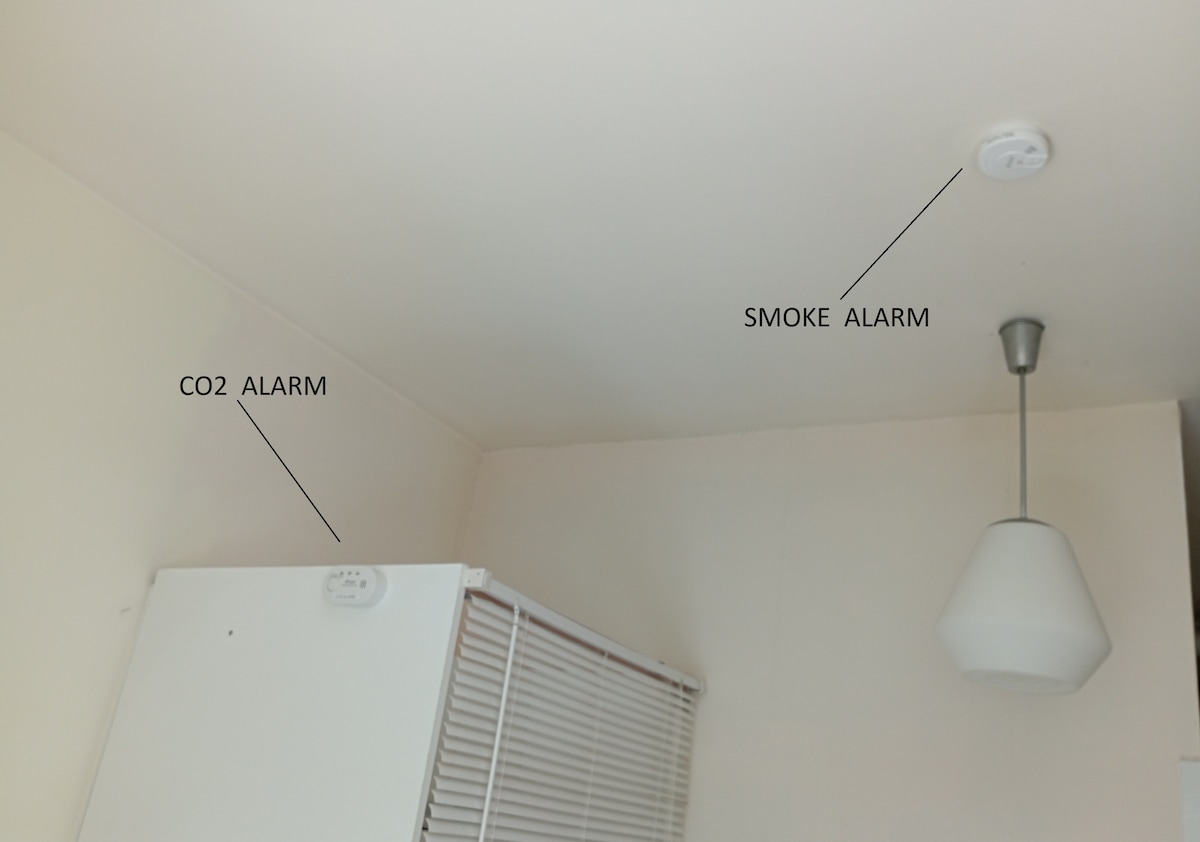 In the image, a smoke alarm and a CO2 alarm are mounted on a light-colored wall near the ceiling. A pendant light hangs from the ceiling, adding functionality to the space. A refrigerator is positioned against the wall, with partially closed blinds visible.