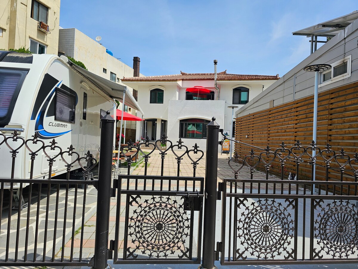 A welcoming entrance is framed by a decorative iron gate, leading to a spacious courtyard. The area is lined with modern structures, featuring a blend of vibrant red umbrellas and wooden accents, allowing for a relaxed outdoor atmosphere.