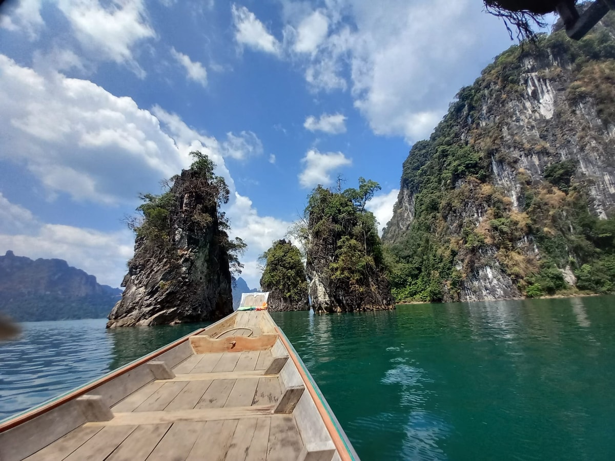 A long-tail boat is anchored on calm turquoise water, surrounded by lush green cliffs and rocky formations. Towering limestone cliffs rise dramatically on either side, with patches of greenery clinging to their surfaces. The sky above is partly cloudy, creating a serene atmosphere.