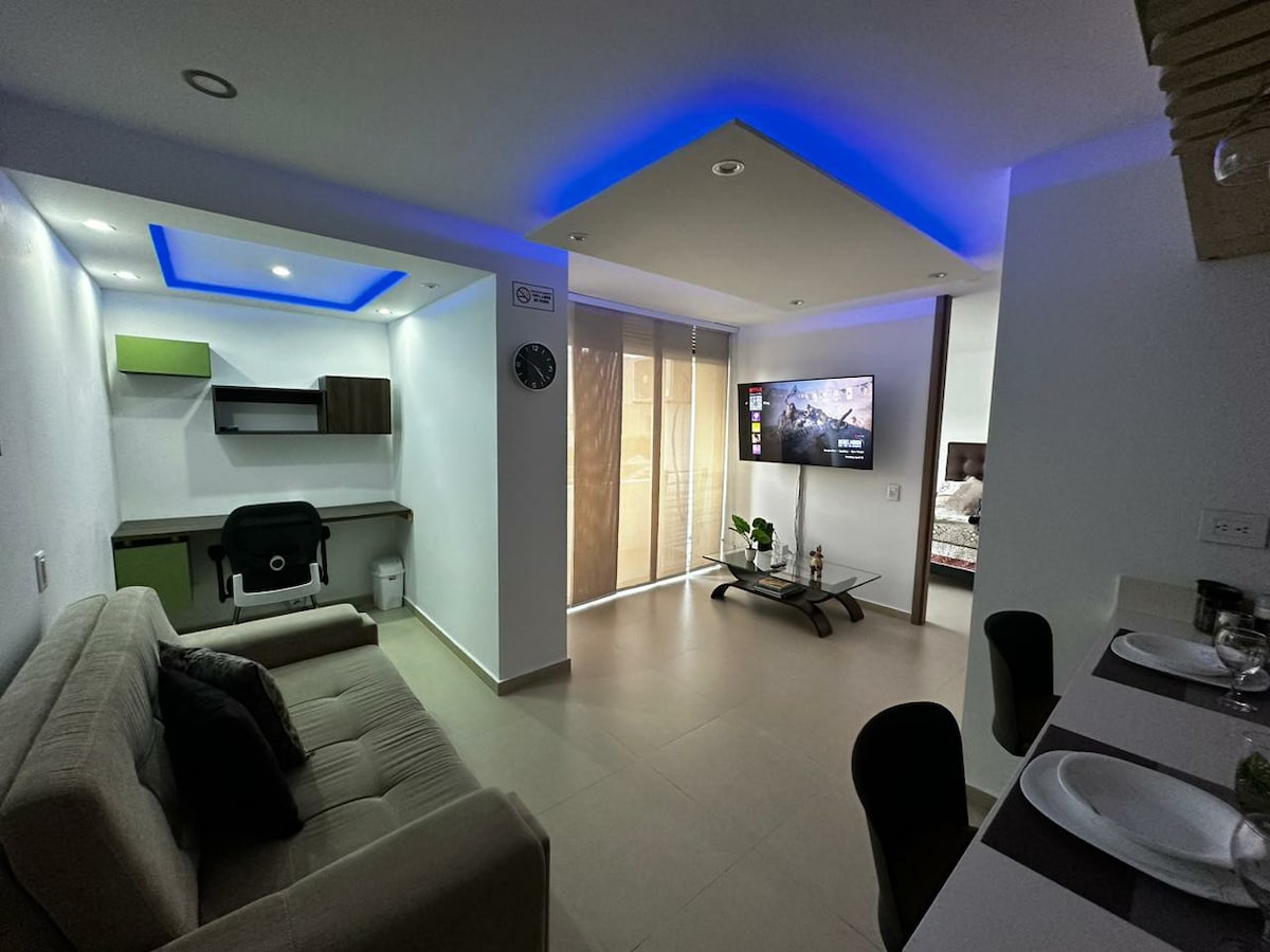A modern living area features a comfortable sofa and a sleek television mounted on the wall. Ambient lighting creates a calming atmosphere. A dining area is visible with minimalist table settings. Large windows allow natural light, with partially translucent coverings providing privacy.