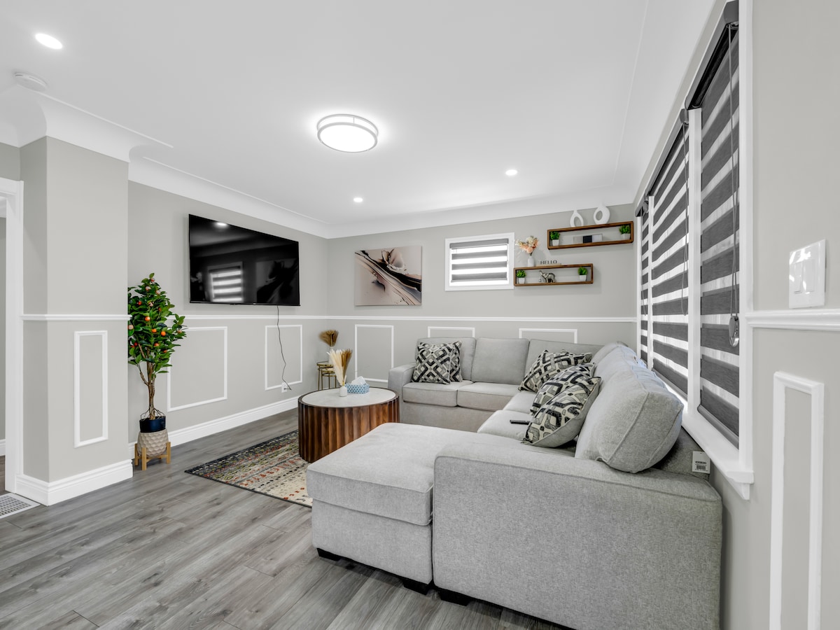 A modern living area features a light gray sectional sofa paired with decorative pillows. A round wooden coffee table is positioned in front, while a wall-mounted TV and shelving with plants add visual interest. Natural light enters through stylish window treatments.