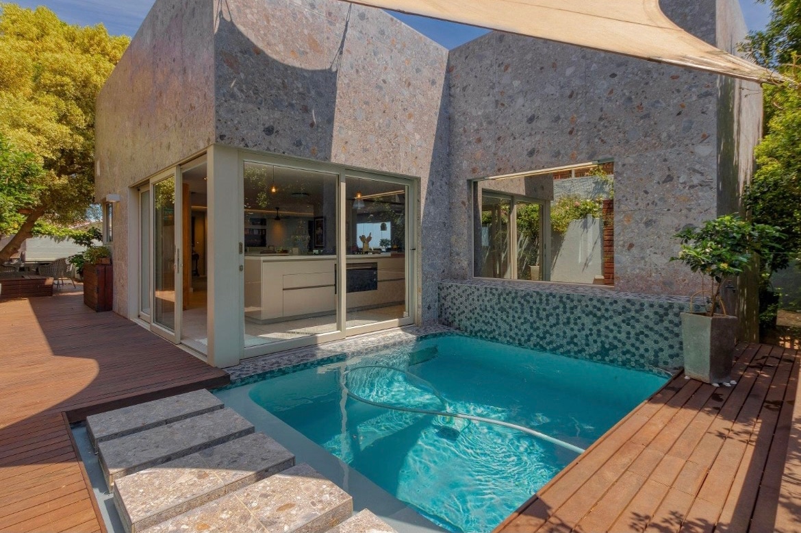 A modern retreat features a sleek pool bordered by light wooden decking. Large glass panels offer views of the interior space, highlighting a spacious kitchen area. The stone exterior blends harmoniously with surrounding greenery, creating a serene outdoor setting.