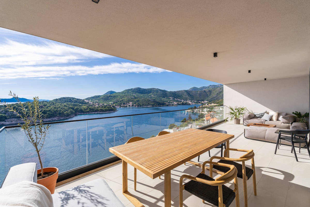 A spacious covered terrace features a wooden dining table surrounded by four chairs. Comfortable seating areas are arranged along the side, with expansive views of the sea and hills visible through large glass panels.