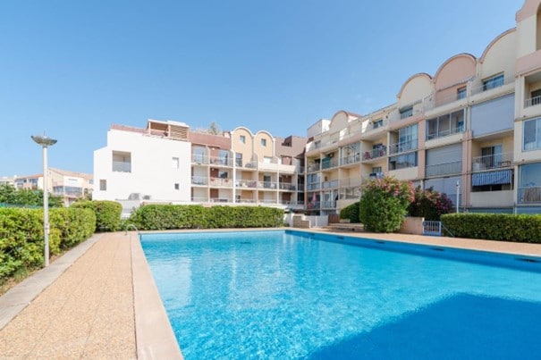 A large swimming pool reflects the blue sky, surrounded by well-kept greenery. The pool area is framed by a residential building featuring multiple balconies and access points. Clear pathways lead to the pool, providing a welcoming space for relaxation.