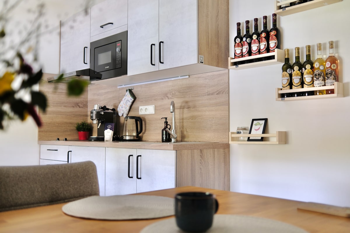 The kitchen area is equipped with modern appliances including a microwave, coffee maker, and kettle. A selection of beverages is neatly displayed on wall-mounted shelves. Wooden cabinets and a dining table create a functional and welcoming space.