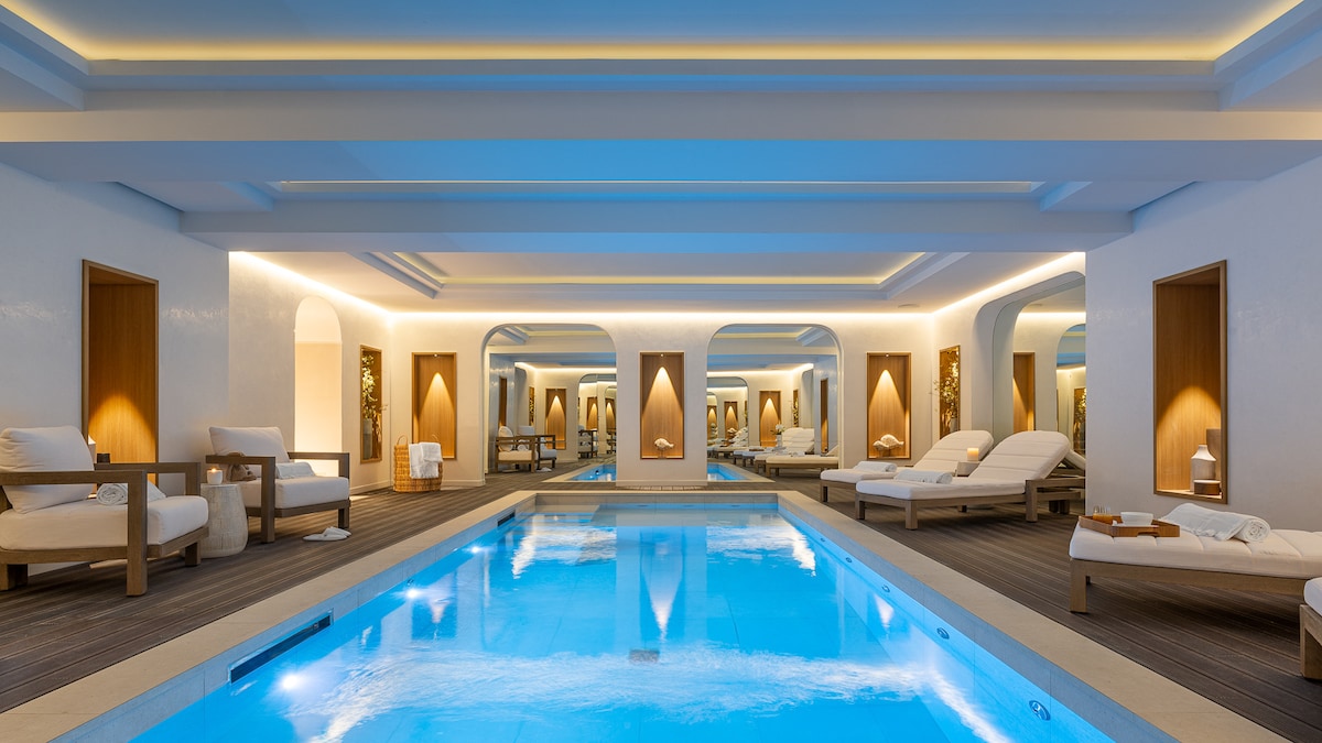 An indoor spa area features a serene swimming pool with clear blue water. Lounge chairs are positioned along the poolside, and elegantly designed archways lead to private treatment rooms, all illuminated by soft, ambient lighting.