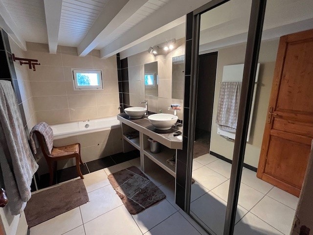 A spacious bathroom features modern fixtures and a double vanity with vessel sinks. A bathtub is positioned against a wall, alongside a wooden chair. Natural light enters through a small window, and the room is adorned with soft towels and textured floor mats.