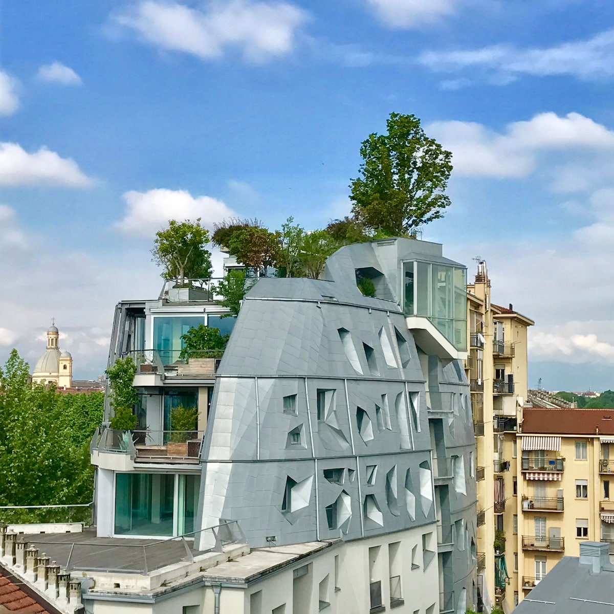 A modern architectural building features a unique faceted design with greenery integrated into its structure. Various window shapes create an interesting facade. The surrounding area includes traditional buildings and trees, contributing to a diverse urban landscape against a partly cloudy sky.