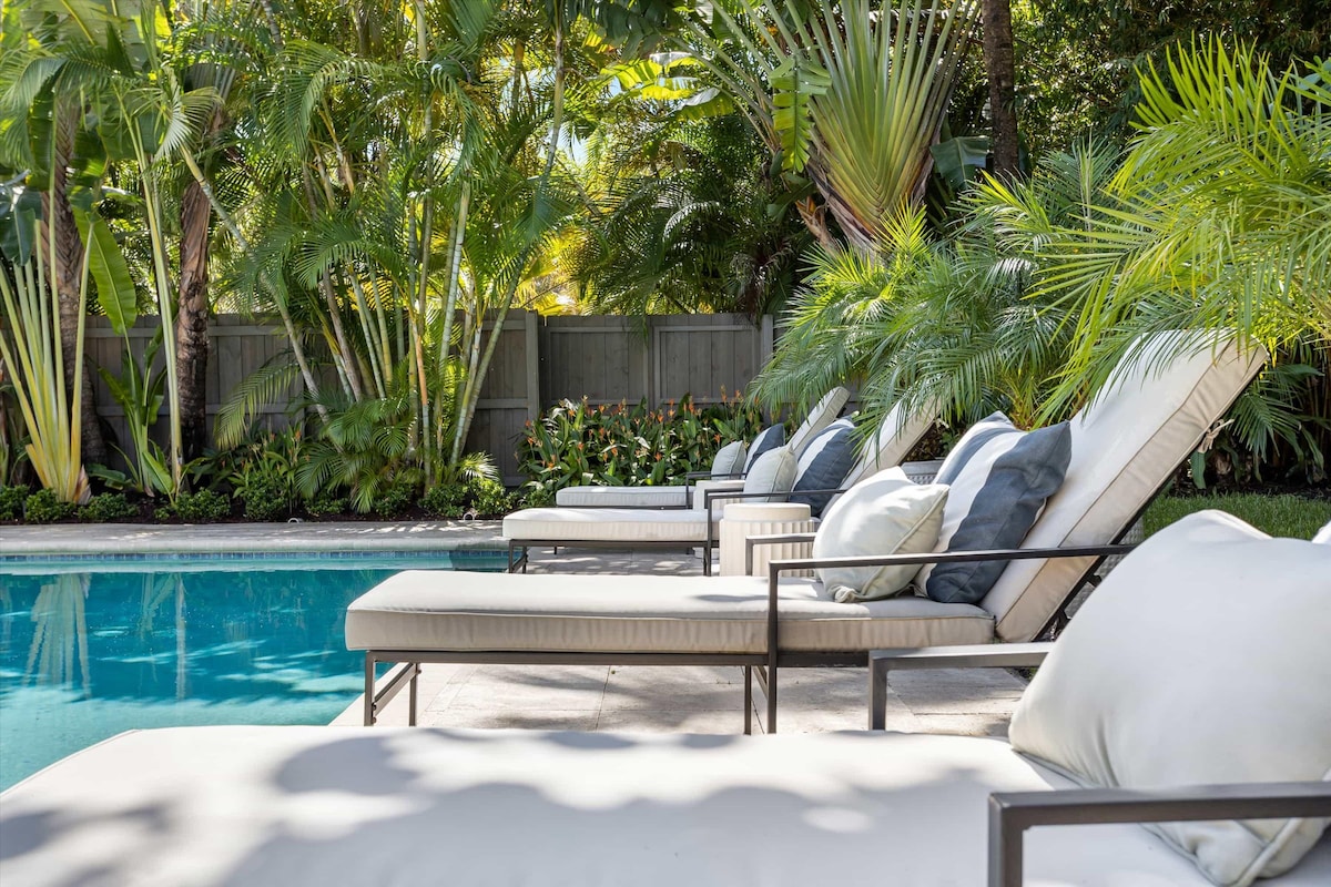 Lounge chairs are positioned beside a tropical heated pool, surrounded by lush greenery and palm trees. Soft cushions are placed on the chairs, creating a relaxing atmosphere. The pool reflects the vibrant foliage, enhancing the space's tranquil appeal.