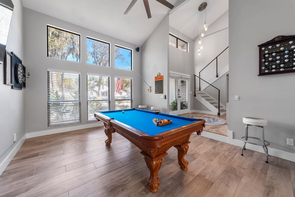 A billiards table with a blue felt surface is positioned in a bright room featuring large windows. Natural light fills the space, highlighting the wooden floor and contemporary fixtures. A staircase leads to the upper level, complementing the modern design of the area.