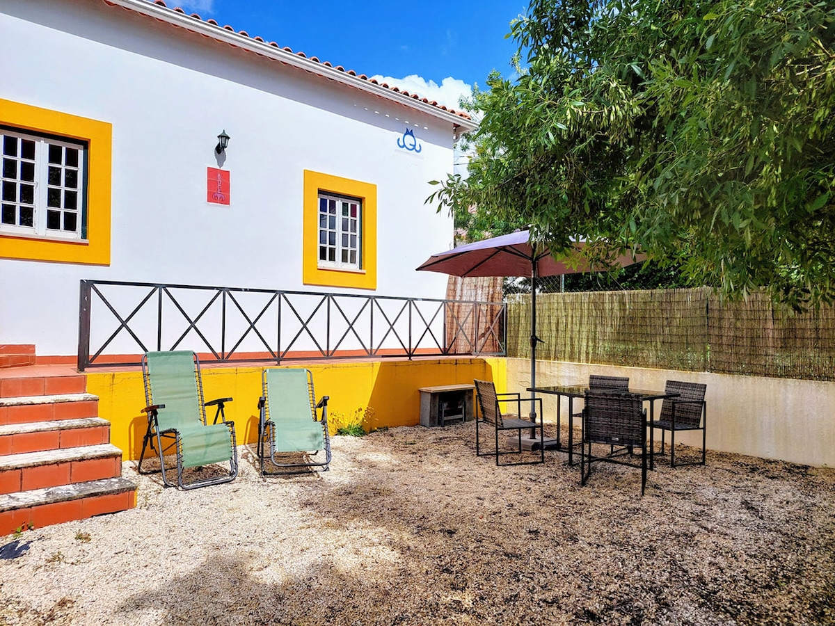 An outdoor seating area is presented, featuring two green lounge chairs and a dining set with four chairs under a shade umbrella. A gravel surface and a nearby fence enhance the secluded feel of the space, with greenery surrounding the area.
