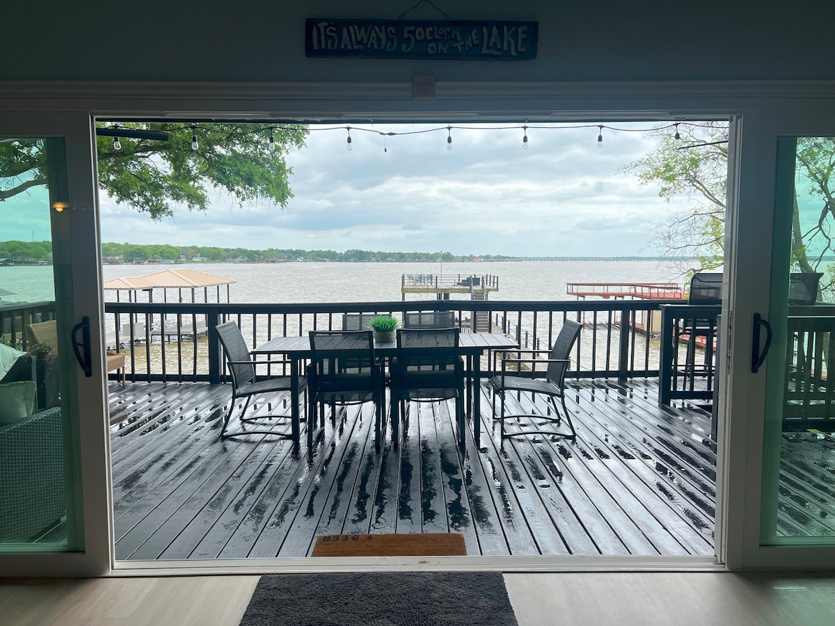 A spacious patio deck is showcased, featuring a dining table with ample seating. Large sliding glass doors highlight views of the tranquil lake and distant docks. The deck surface is composed of weathered wood, and the area is adorned with string lights, providing a cozy atmosphere.