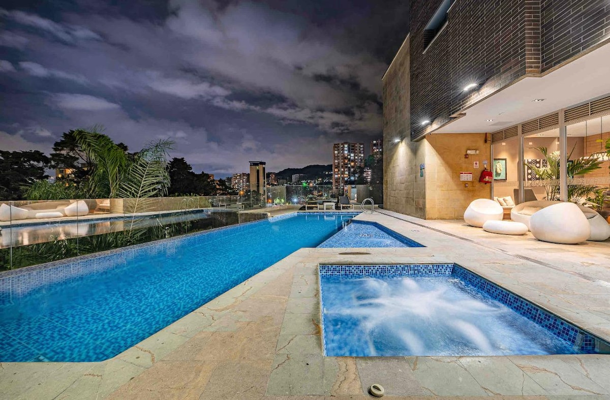 An inviting swimming pool area is illuminated under the night sky, featuring a large, clear pool alongside a heated jacuzzi. Lush greenery and modern seating create a tranquil atmosphere, while city lights are visible in the background, enhancing the serene environment.