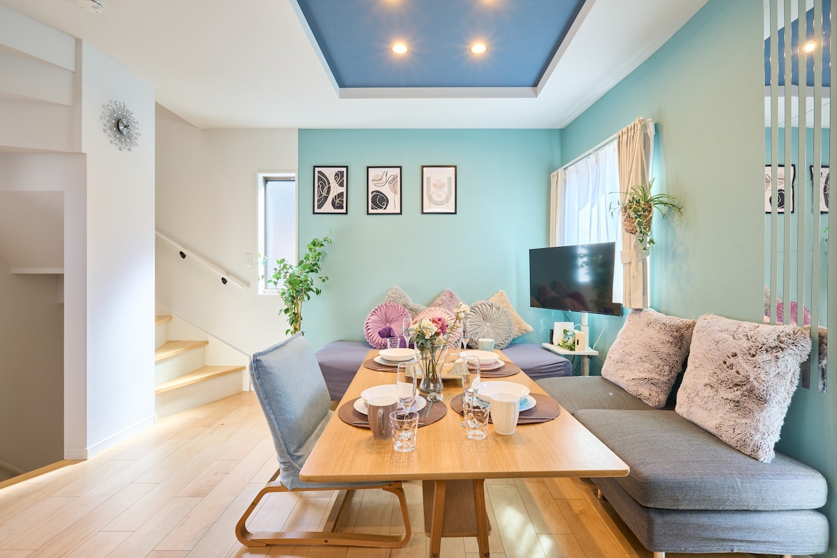 A dining area with a wooden table is set for a meal, displaying dishes and glasses. A cozy seating area features soft cushions next to a bed. Natural light enters through the window, illuminating the blue accent ceiling and pastel green walls.