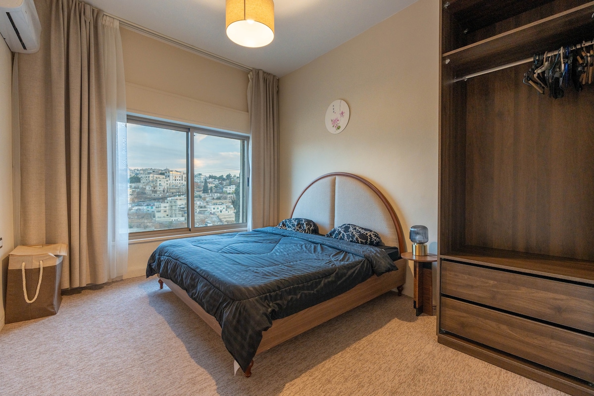 A spacious bedroom is illuminated by natural light from large windows, showcasing views of the surrounding landscape. A generously sized bed with a dark duvet is positioned against a modern headboard. A sleek wooden wardrobe and storage unit are visible, contributing to the room's contemporary aesthetic.