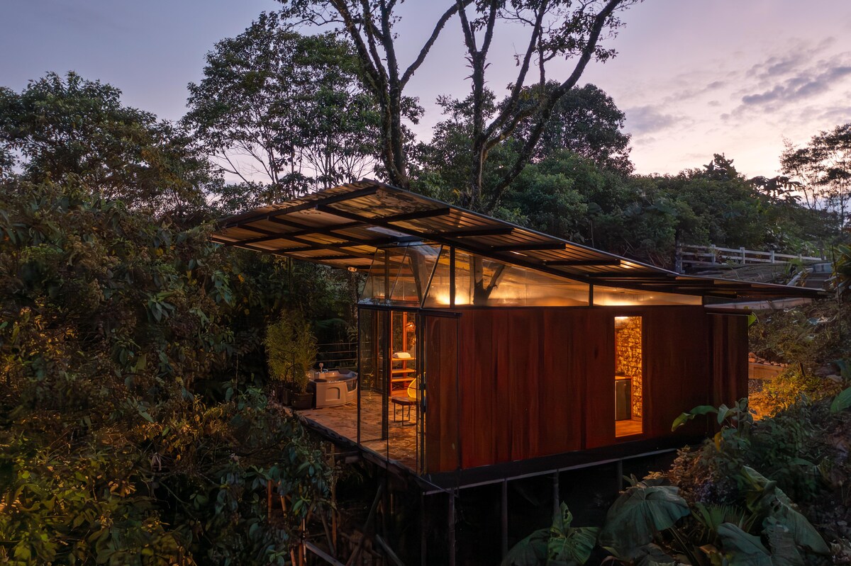 A modern glamping structure is nestled among lush greenery, featuring large glass windows that provide views of the surrounding trees. The roof extends outward, creating a covered outdoor seating area, while the earthy tones of the walls blend harmoniously with the natural landscape.