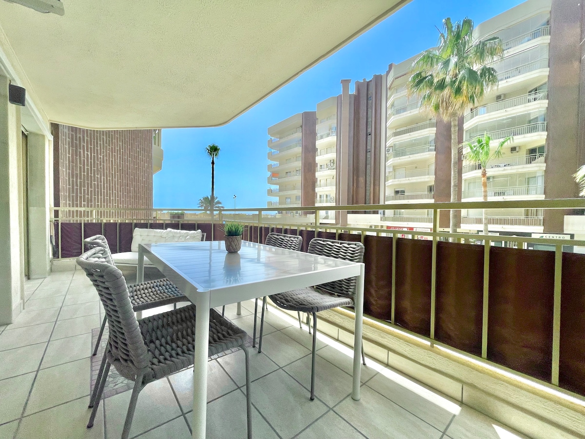 An outdoor terrace is equipped with a rectangular table and several wicker chairs, offering a comfortable setting for dining al fresco. Palm trees and nearby buildings are visible, providing a glimpse of the vibrant surroundings.