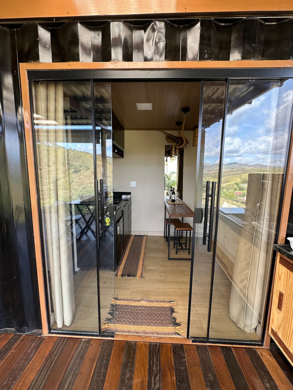 An entryway showcases glass sliding doors leading into a modern kitchen space. High ceilings and wooden accents provide a spacious feel. A dining table with chairs is positioned beside a window, allowing natural light to illuminate the area. Scenic views of the hillside are visible through the glass.