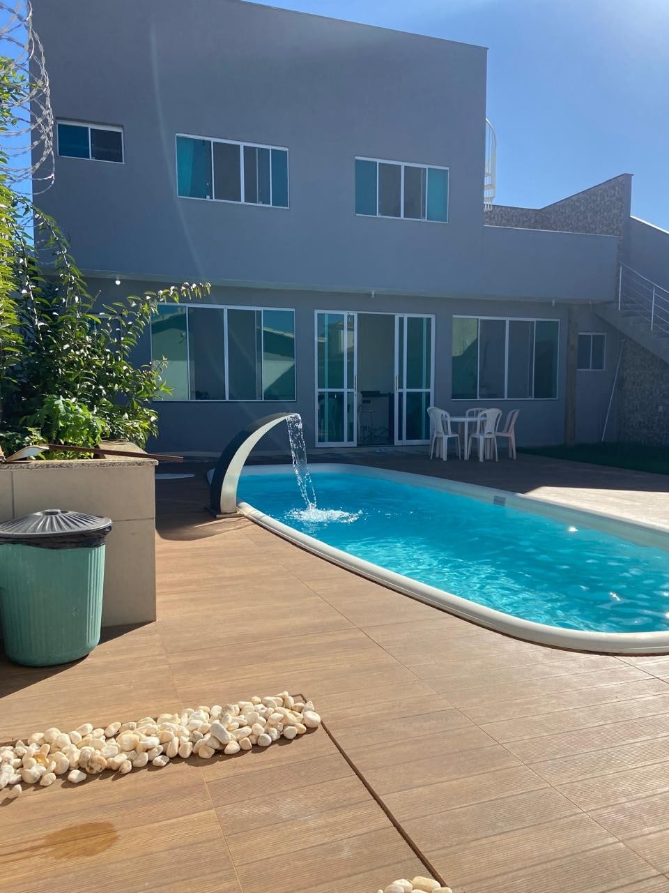 A modern house exterior showcases a tranquil swimming pool featuring a cascading water feature. The pool area is complemented by a wooden deck with decorative pebbles. The light-filled indoor space is visible through large windows, enhancing the connection with the outdoor setting.