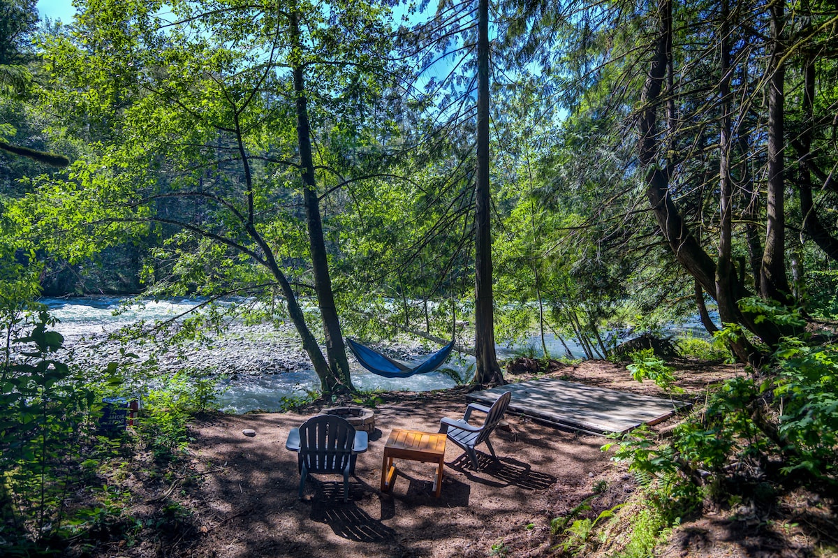 An inviting riverbank area is framed by tall trees, featuring a wooden platform and a small table. Two Adirondack chairs are positioned around a fire pit, while a hammock hangs between the trees nearby. The tranquil river flows gently in the background.