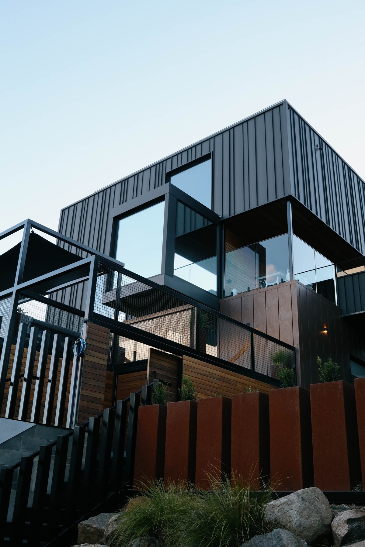 A modern three-story house features a sleek metal exterior with large glass windows, allowing natural light to fill the interiors. The entrance is approached via a staircase surrounded by a blend of wooden and metal elements, contributing to the home's contemporary design.