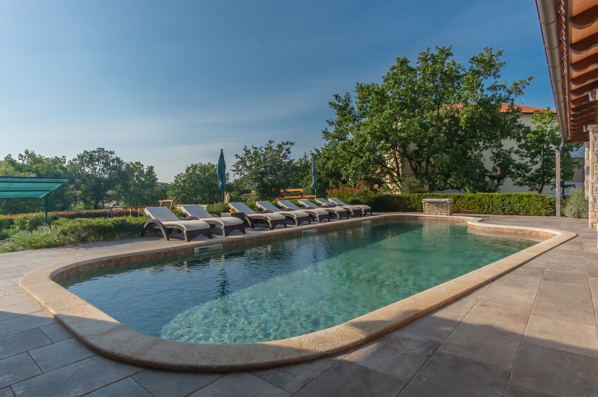 A tranquil private swimming pool stretches across the outdoor area, surrounded by sun loungers and lush greenery. Trees offer shade along the edge, while the clear water reflects the blue sky, enhancing the serene atmosphere of the villa's outdoor space.