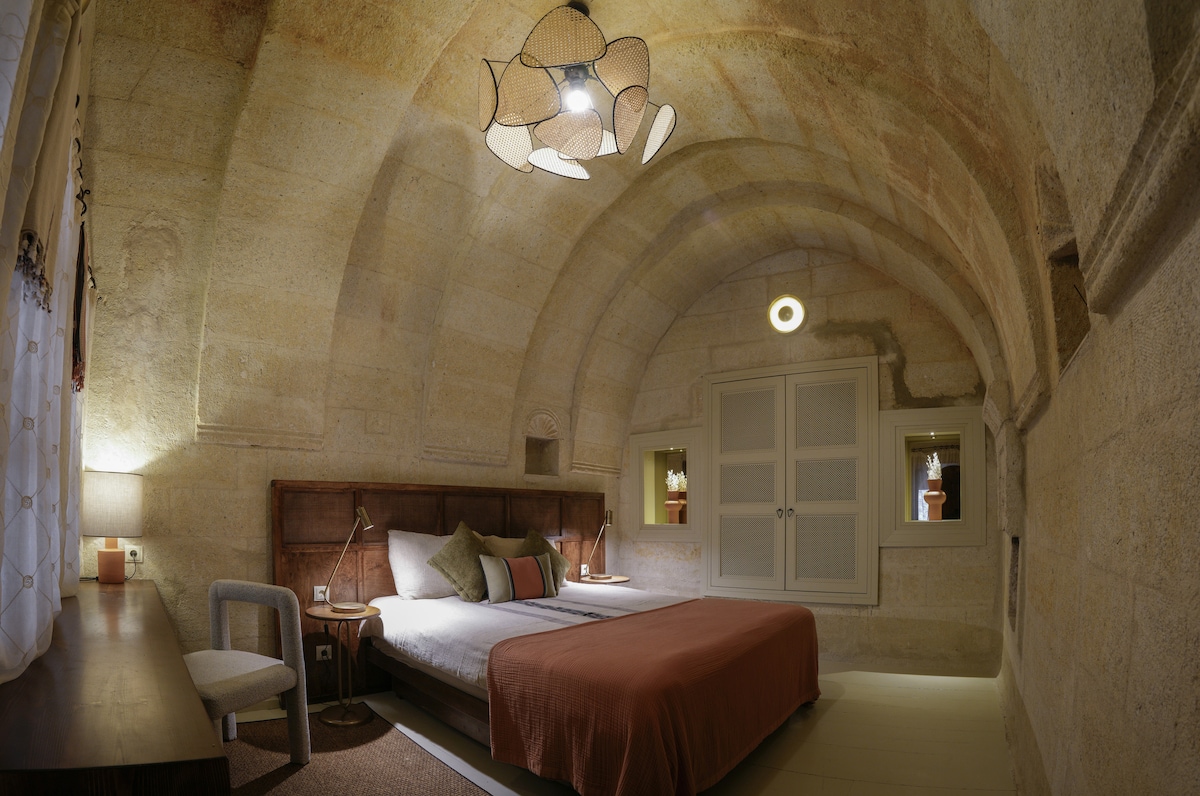 A softly lit room showcases a spacious bed adorned with neutral linens and a rust-colored throw. Arched stone walls create a unique architectural feature, while tasteful decor is highlighted by built-in shelves. A modern chandelier and a simple chair complete the inviting atmosphere.