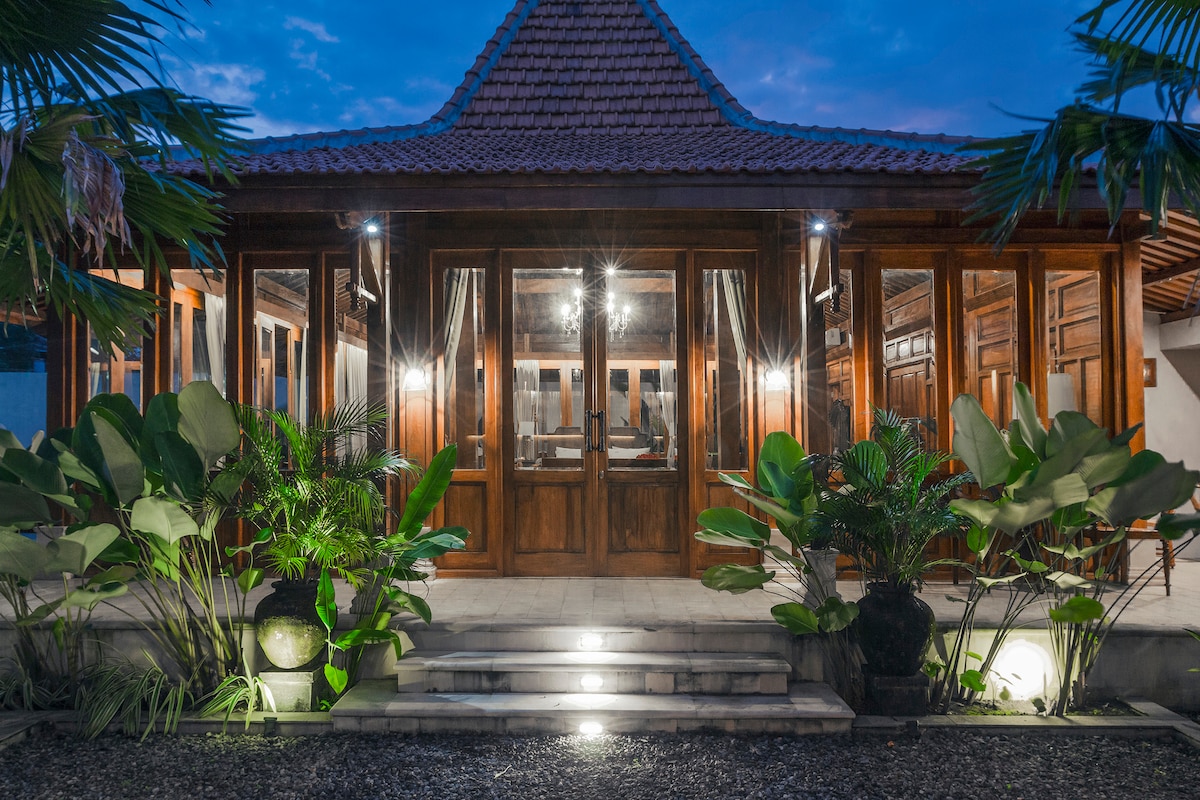 The villa features a traditional joglo design, with large glass-panel doors set in a wooden structure. Lush greenery surrounds the entrance, and soft lighting accentuates the inviting atmosphere as the dusk sky adds warmth to the scene.