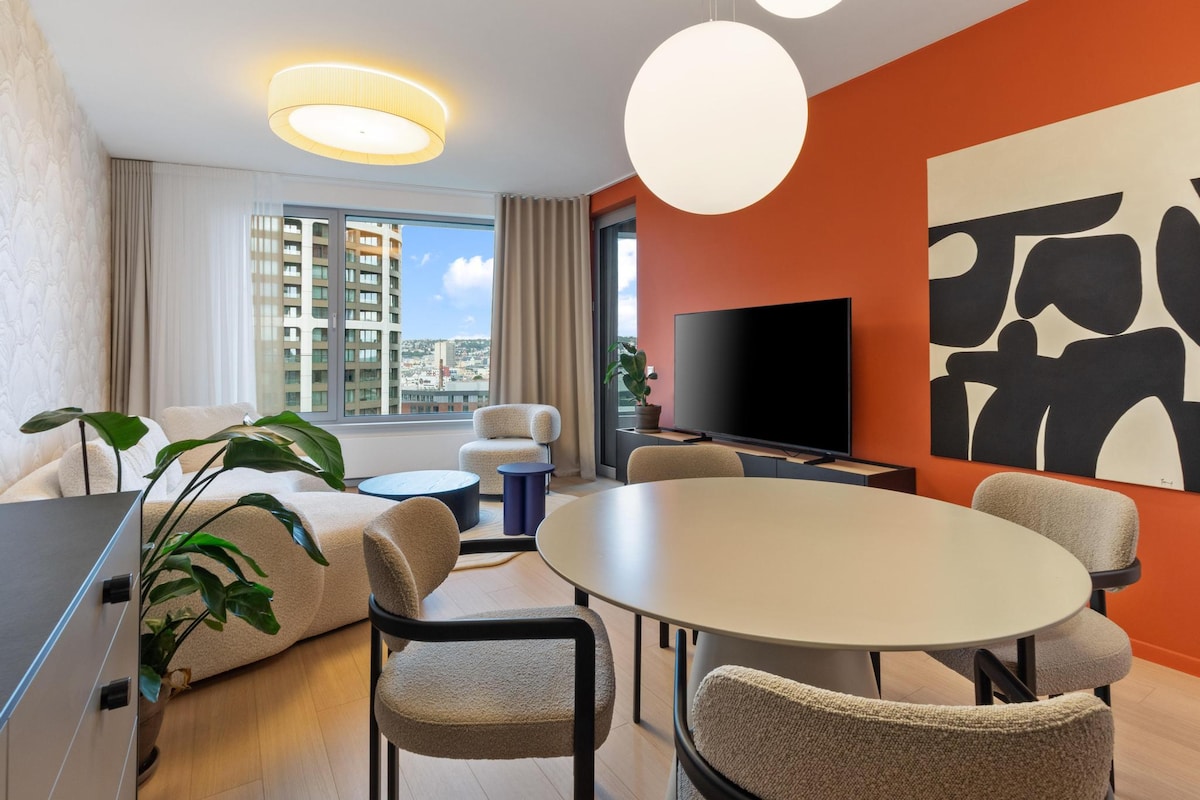 A modern living space is presented with an orange accent wall and a large window offering city views. A light-colored sectional sofa complements a round dining table surrounded by four stylish chairs. A TV is mounted on the wall, and a potted plant adds a touch of greenery.
