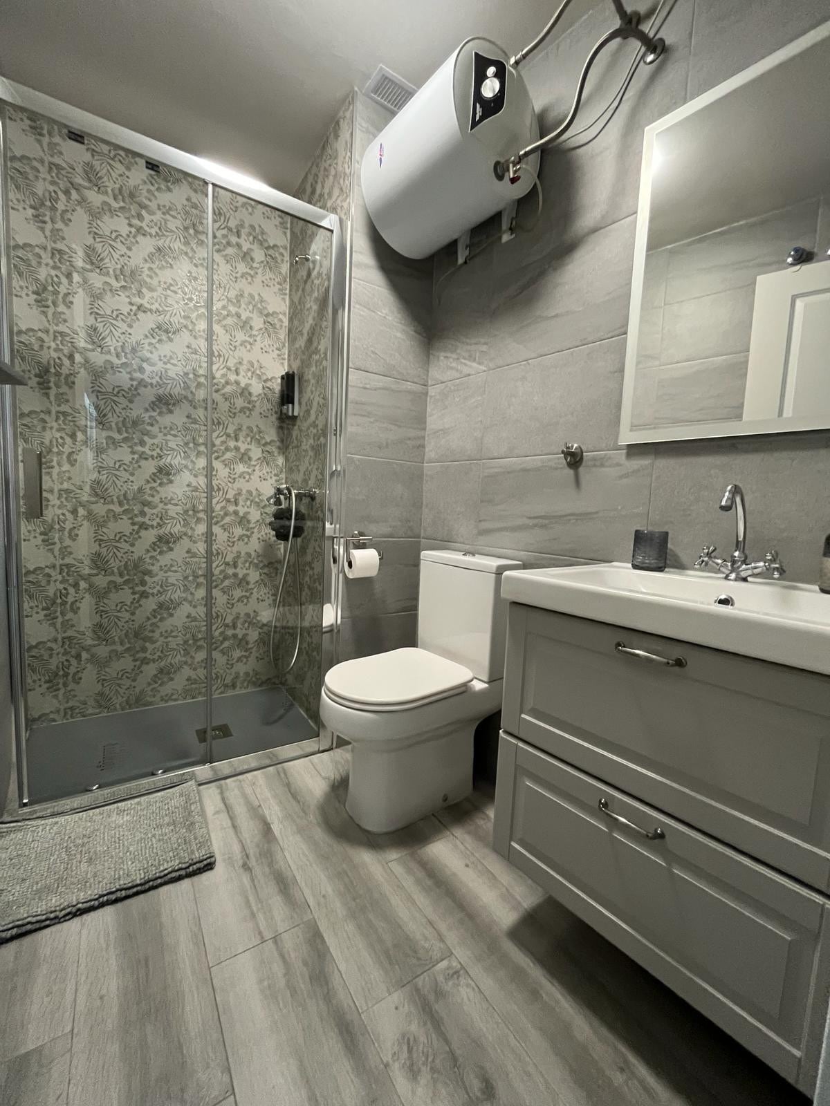 A modern bathroom is presented with a glass shower featuring decorative tiles. A white vanity with storage space is positioned beside a toilet. Neutral tones are complemented by wood-like flooring, while natural light is available through a window obscured by a frosted treatment.