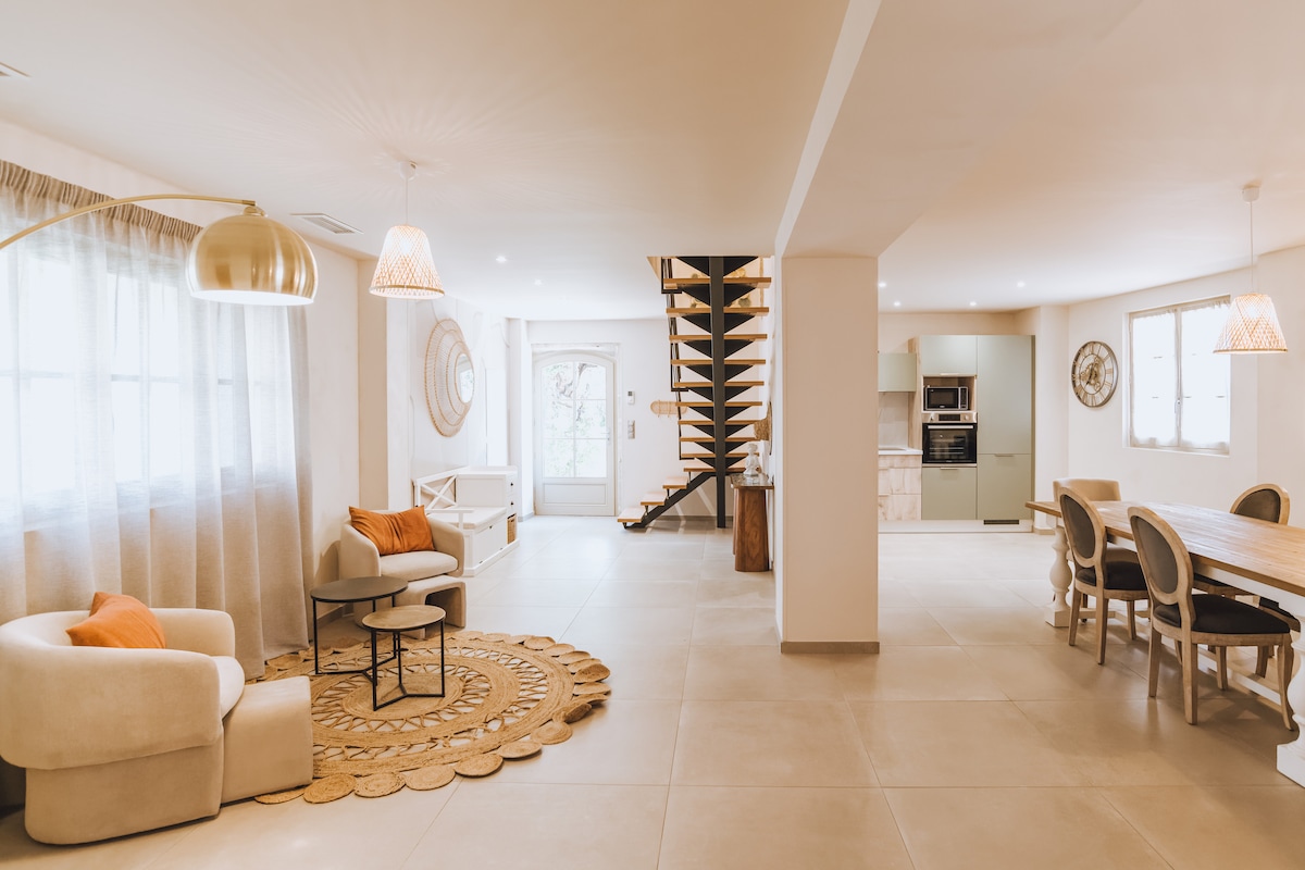The open living area showcases a welcoming design with modern furniture, including soft seating and a wooden dining table. Natural light filters through large windows, illuminating the space. A staircase leads to the upper level, while a round rug adds a touch of warmth to the floor.