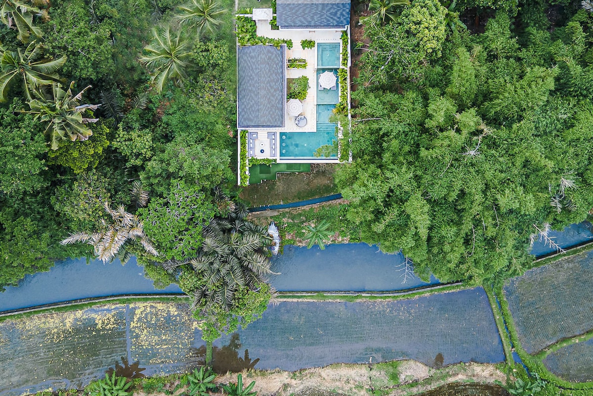 An aerial view captures a modern villa surrounded by lush greenery and tranquil water features. The villa's pool reflects the sky, while outdoor seating is visible. The landscape includes rice paddies and a serene waterway, enhancing the tropical setting.