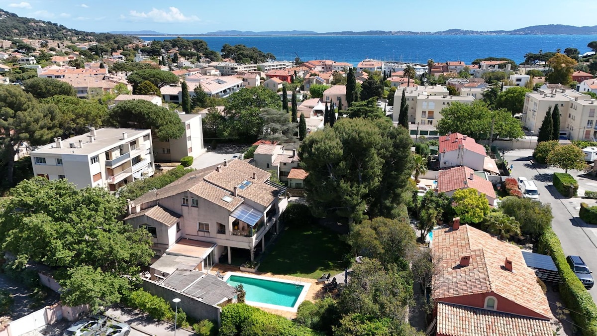 An aerial view captures the peaceful neighborhood of Carqueiranne, featuring the duplex with a pool surrounded by lush greenery. The coastline is visible in the background, with residential buildings lining the streets, while the vibrant tree canopy adds to the area’s tranquility.