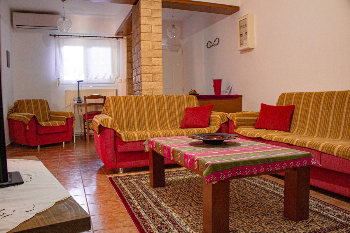 A welcoming living area features two red sofas with striped cushions and wooden chairs. A low wooden table is placed at the center, adorned with a decorative cloth. Natural light enters through a window, illuminating the cozy space with warm tones.