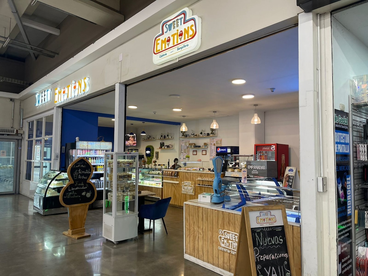 A modern storefront is displayed, featuring a bright blue accent wall and wooden counters. Refrigerators are stocked with various products, and signage is visible overhead. Pendant lights hang from the ceiling, illuminating the shop's interior, where customers can be seen exploring the offerings.