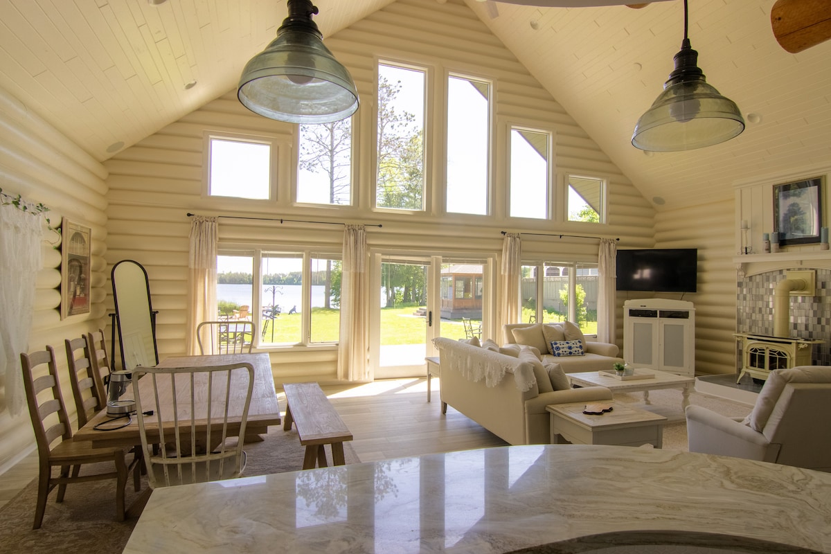 An open-concept living area features high ceilings with exposed beams and large windows allowing for an abundance of natural light. A comfortable seating arrangement is visible alongside a dining table. View of the lake and outdoor space is framed by the expansive glass panels.