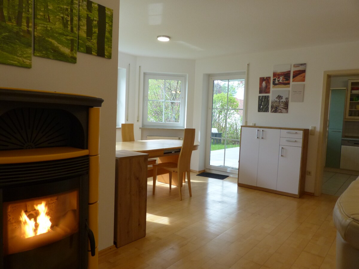 A spacious room features a warm pellet stove, complemented by a wooden dining table surrounded by chairs. Large windows allow natural light to permeate the area, showcasing a serene atmosphere. Artwork is displayed on the walls, enhancing the room's inviting appearance.