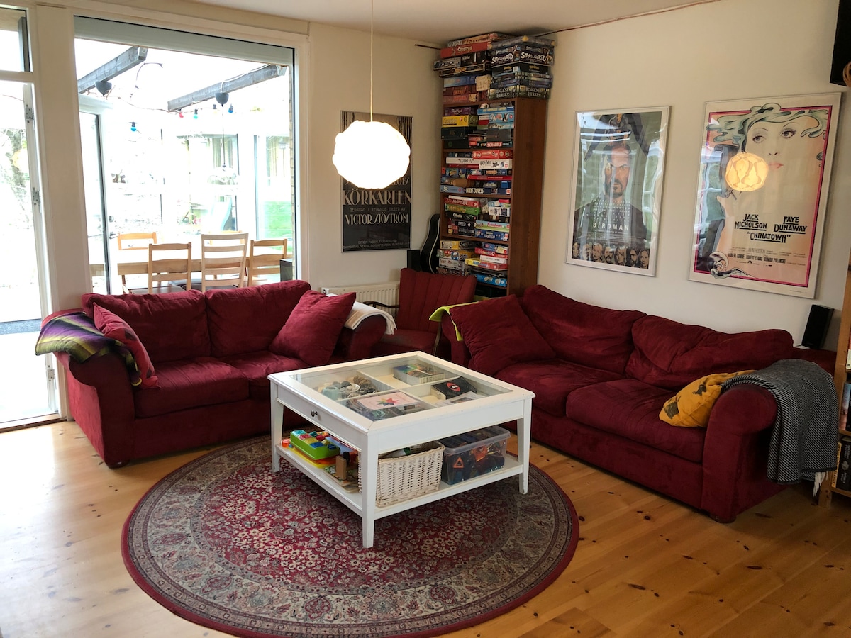 A cozy living area features two red sofas arranged around a white coffee table, which holds various games and books. A large window allows natural light to fill the space, with a dining area visible in the background. A decorative rug adds warmth to the wooden floor.