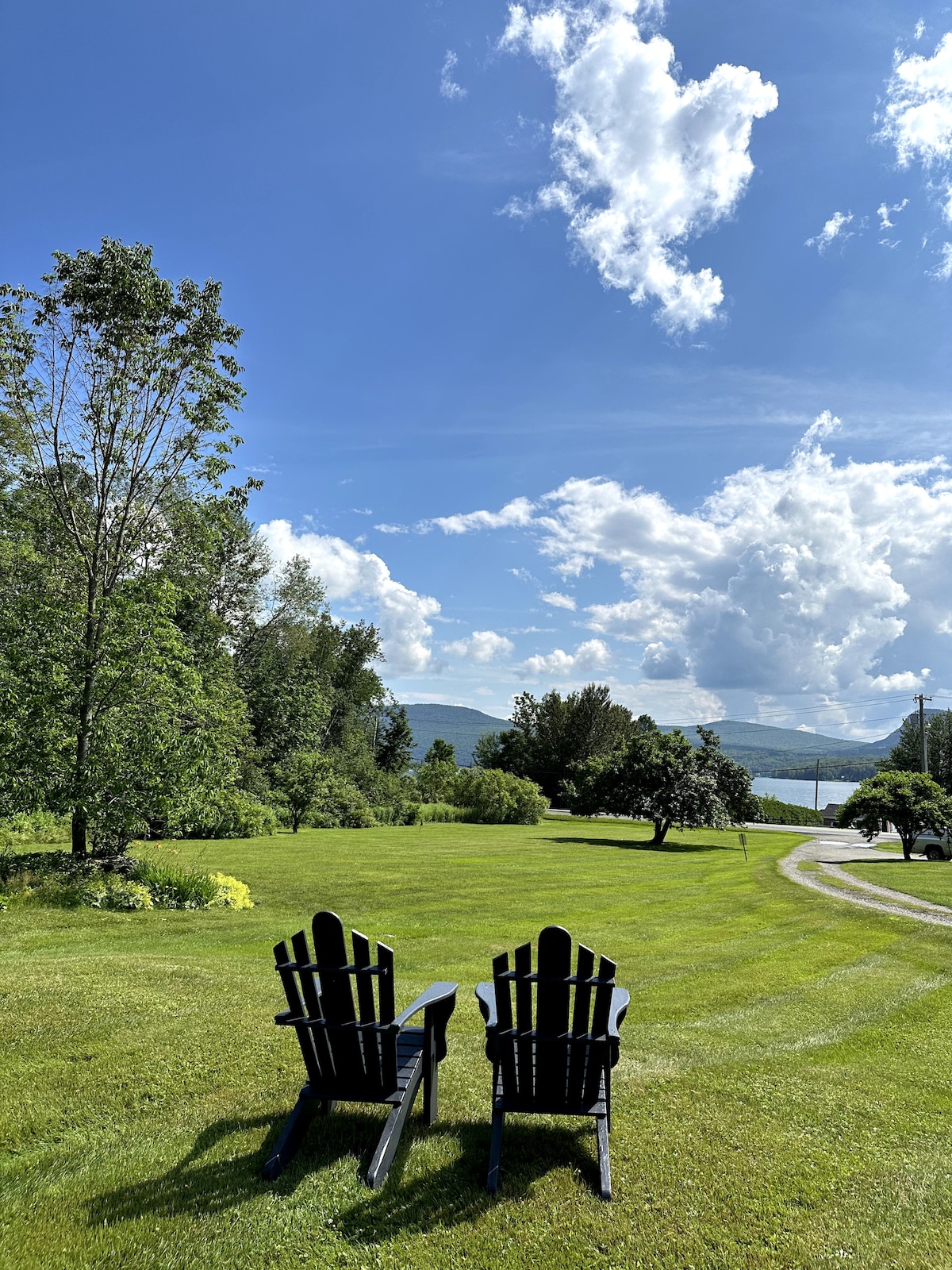 Two black Adirondack chairs are positioned on a lush green lawn, overlooking a scenic view of distant mountains under a partly cloudy sky. The area is surrounded by trees and well-maintained landscaping, providing a serene outdoor space.