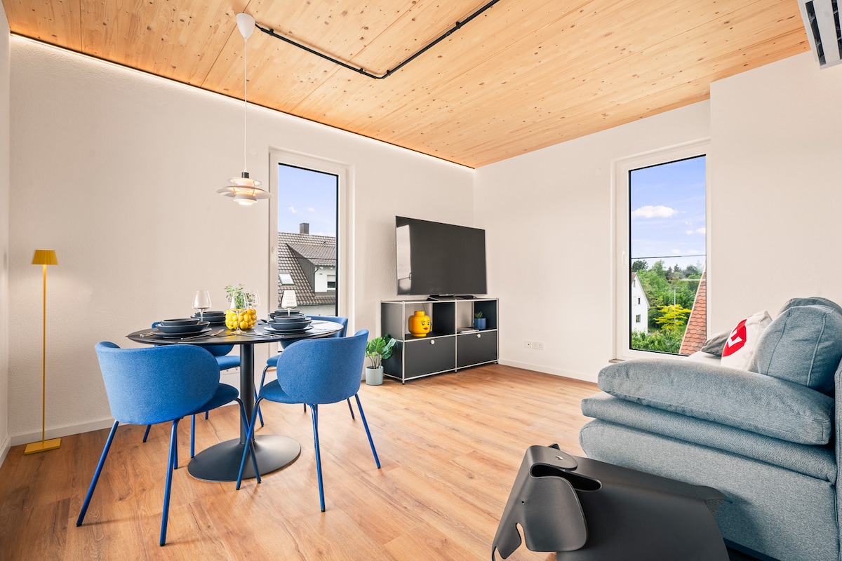 A contemporary living area features a round dining table set for four, paired with vibrant blue chairs. A flat-screen television is positioned on a sleek black unit against a wall of light. Two windows allow natural light to fill the space, highlighting wooden flooring.