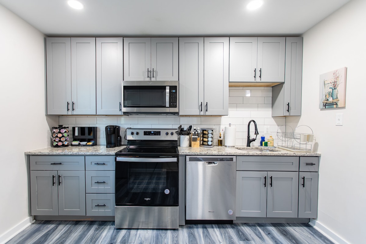 A modern kitchen setup is displayed, featuring gray cabinetry and granite countertops. Stainless steel appliances, including a microwave, oven, and dishwasher, are visible. A coffee maker and kitchen essentials are neatly organized on the counter, enhancing the functionality of the space.