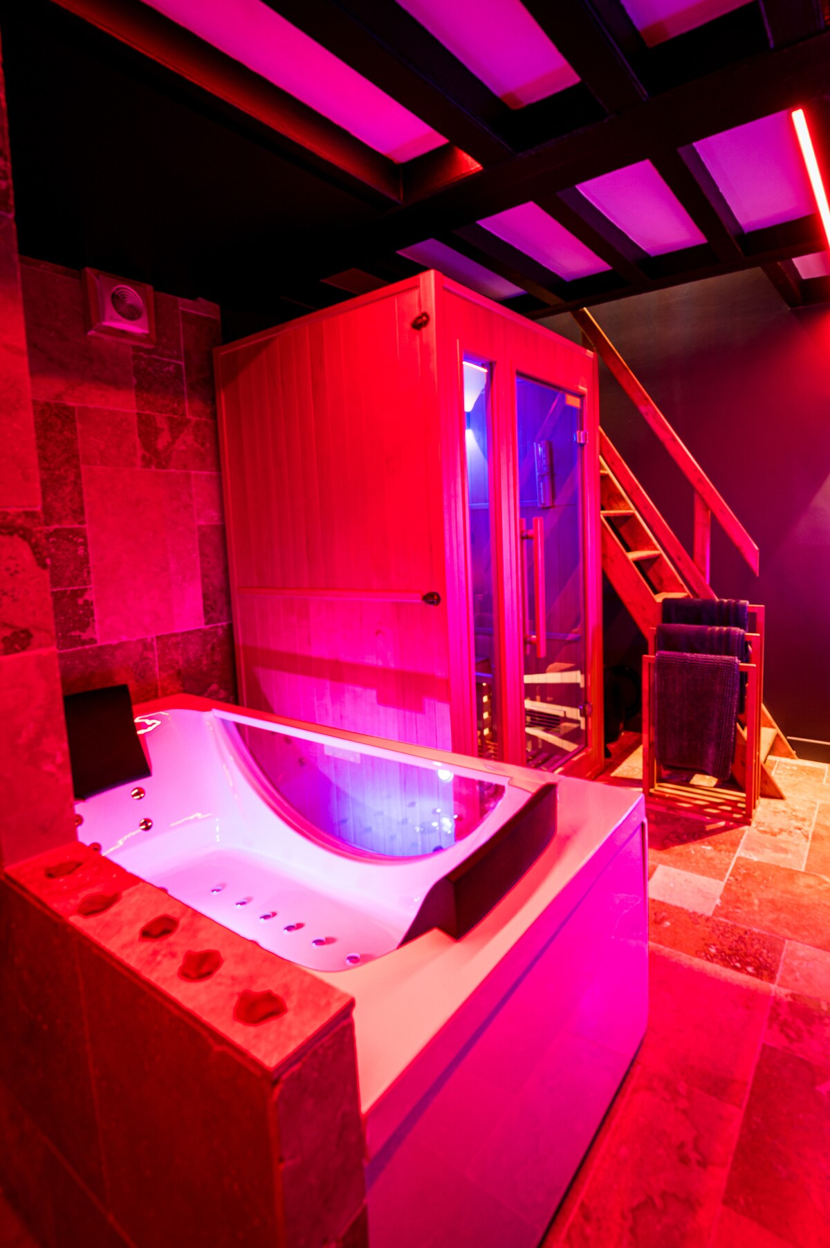 A wellness area is highlighted, featuring a whirlpool bathtub with ambient lighting. A wooden sauna is visible alongside, with a staircase leading to a separate space. Textured stone walls enhance the unique atmosphere, complemented by soft pink and purple lighting.