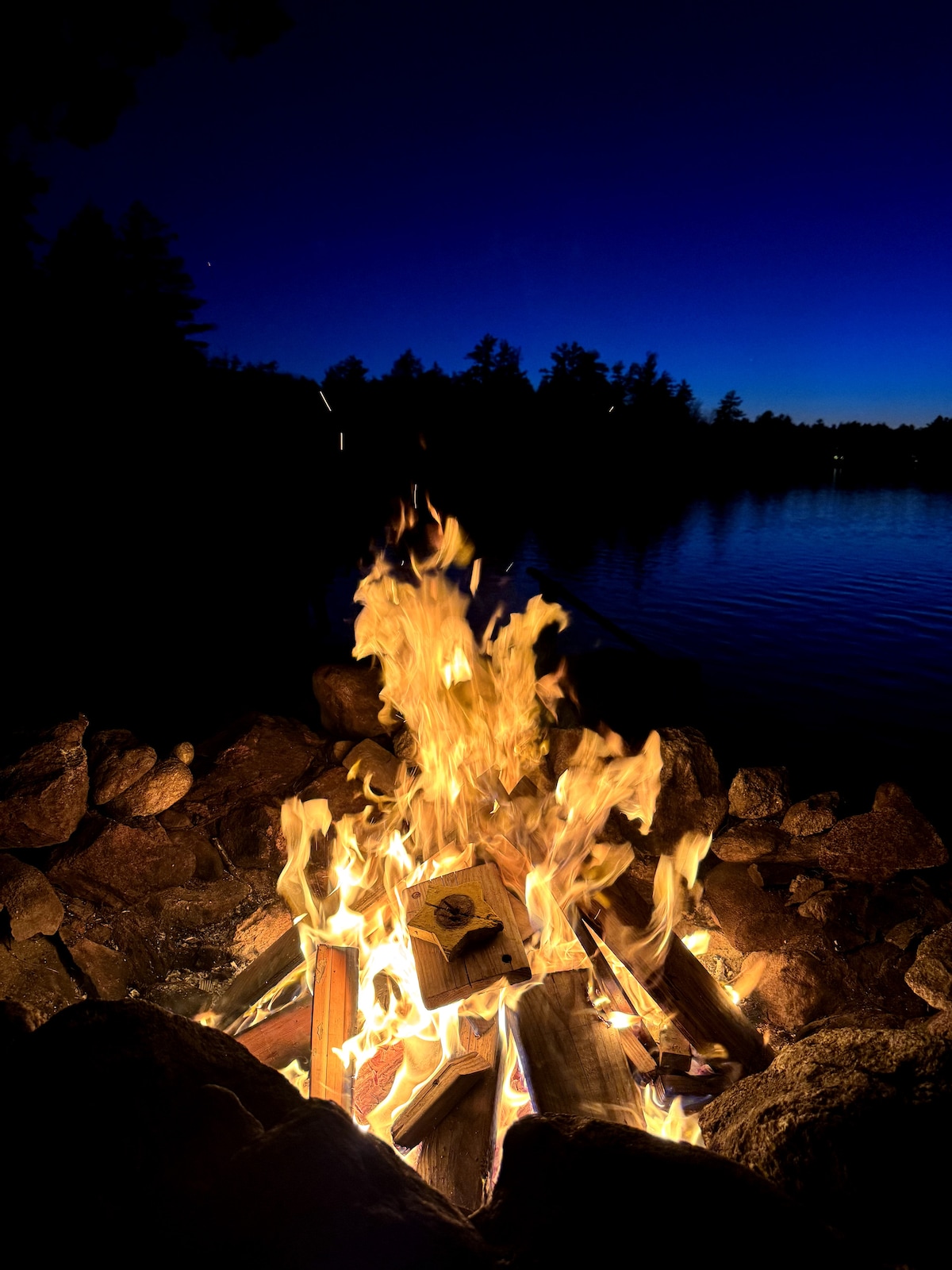 A crackling campfire is framed by a circle of smooth stones, casting a warm glow against the approaching twilight. Reflections of the fire dance on the nearby lake, creating a serene atmosphere amidst the surrounding trees.
