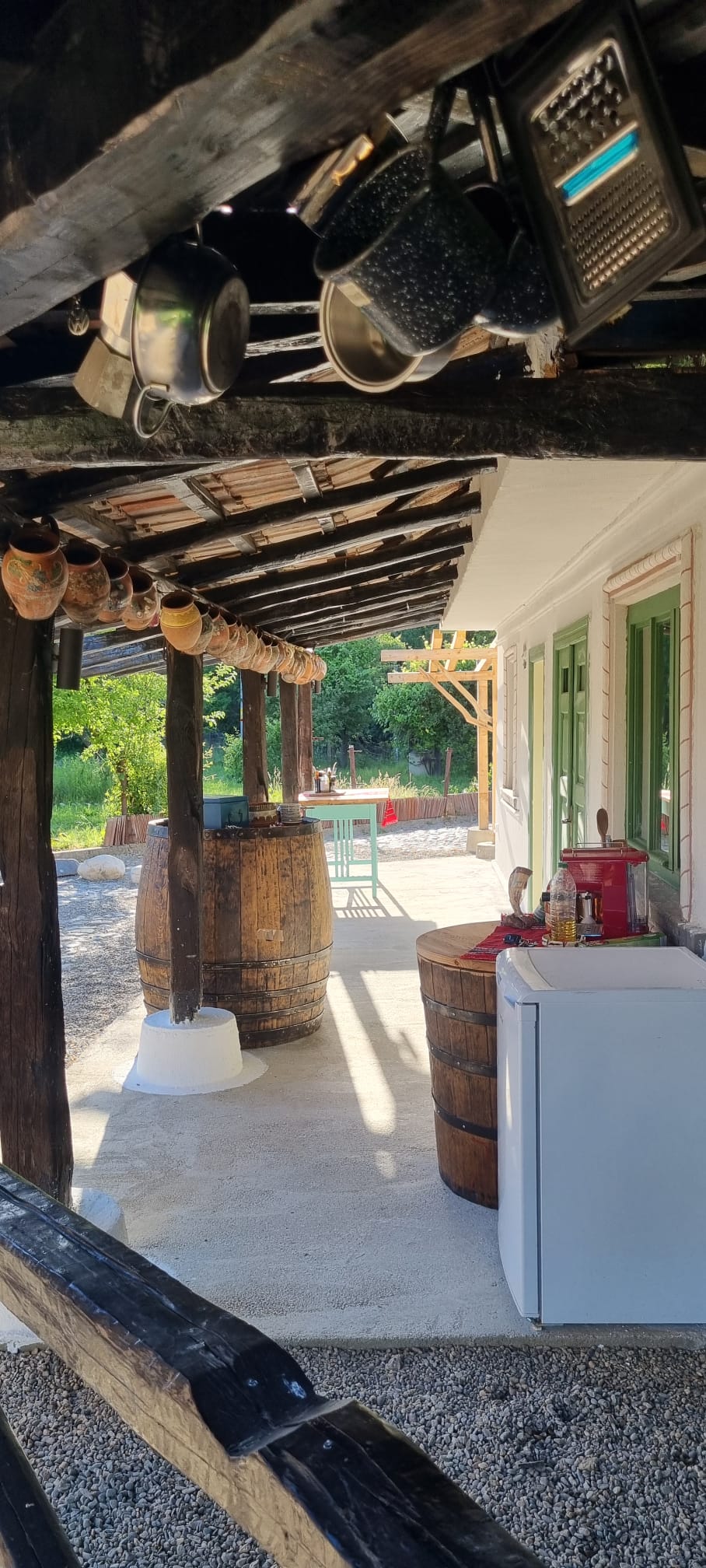 An outdoor kitchen area is featured, showcasing wooden beams overhead and various kitchen items hanging above. Barrels serve as decorative elements, alongside a small refrigerator and a red appliance on a table. Natural light illuminates the space, creating a welcoming environment.