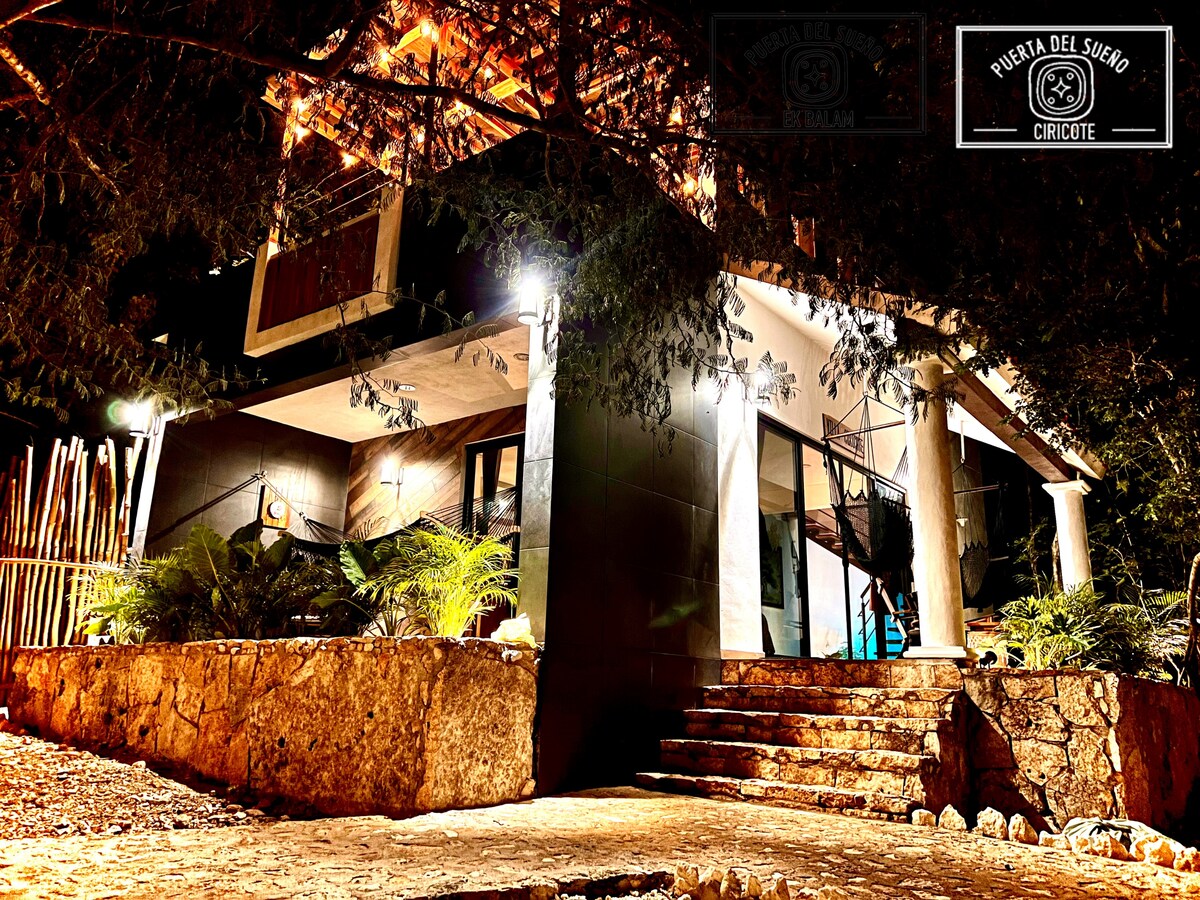 The exterior of a modern property is shown, featuring a two-story design with a well-lit entryway. Natural stone steps lead to a spacious porch, adorned with tropical plants and illuminated by soft outdoor lighting at night.