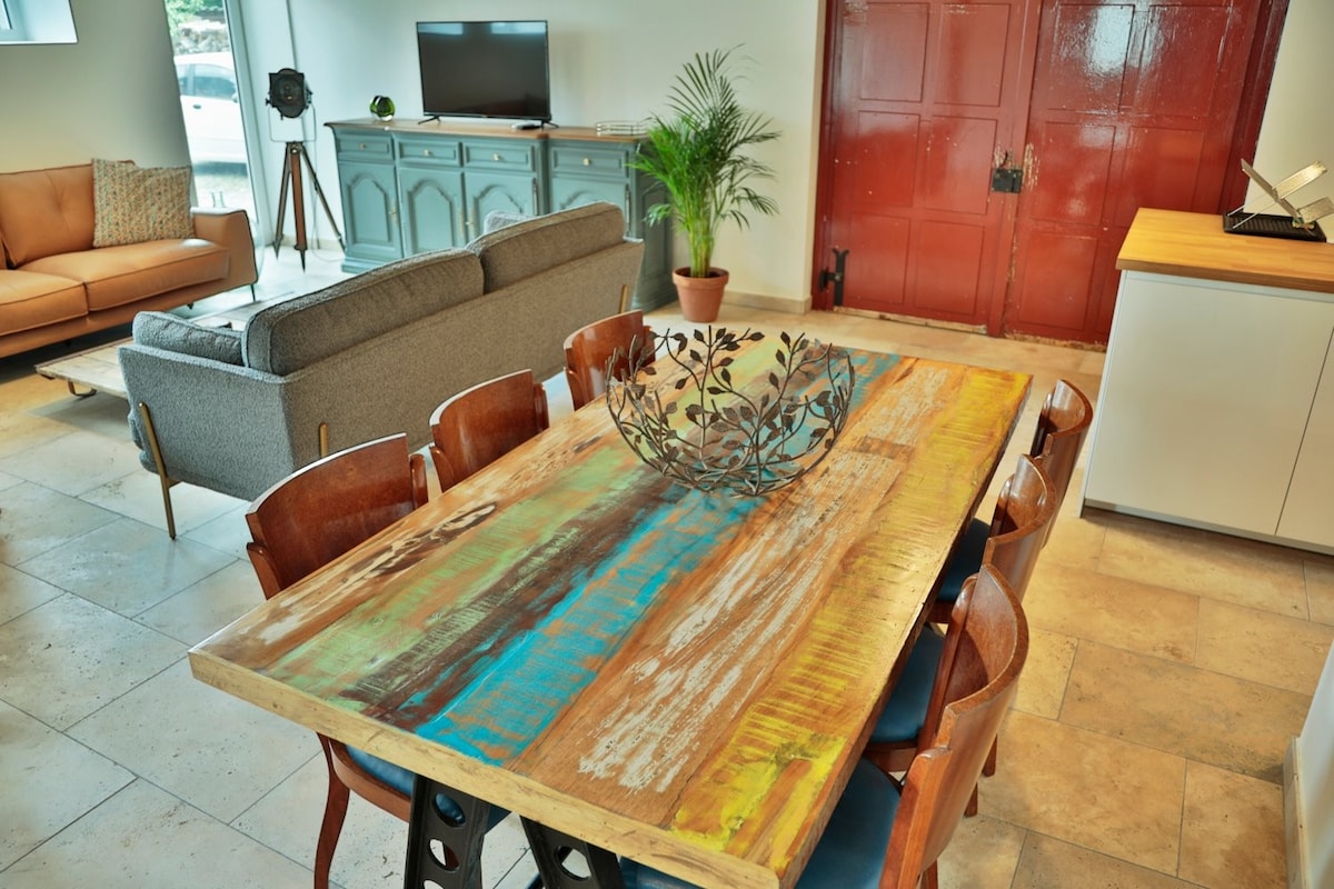 A dining area features a large wooden table with a colorful surface, surrounded by several wooden chairs. A decorative metal centerpiece adds interest. The open layout connects to a comfortable seating area and a television set against a backdrop of red doors.